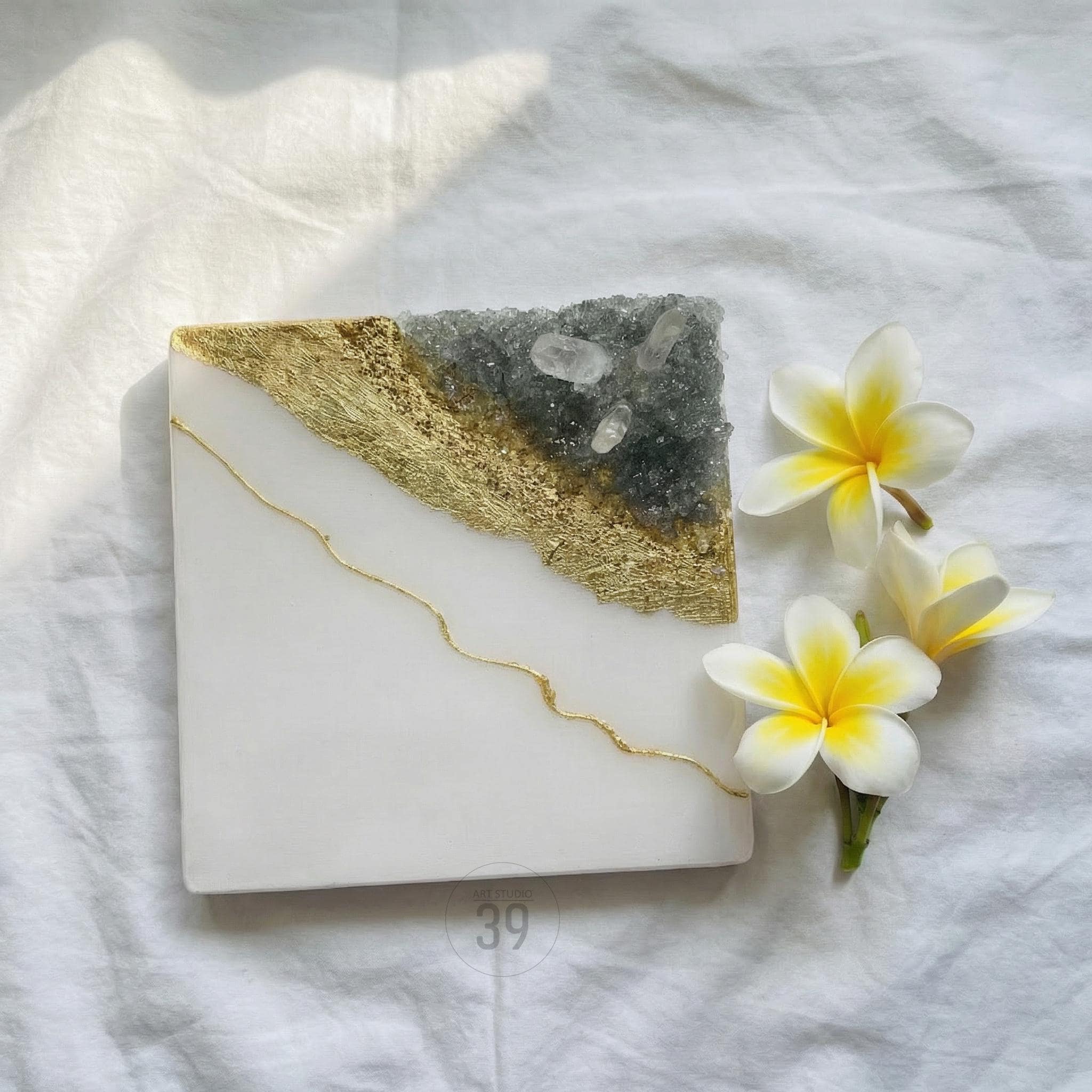 Square white resin coaster with a gold leaf and gray crystal design. A gold line accents the white surface. Three white and yellow flowers are placed on the right side of the coaster.