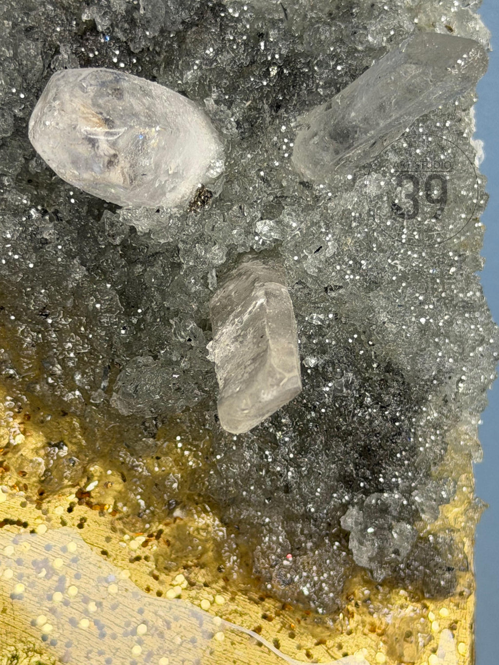 Close-up of a mineral specimen featuring clear, crystalline formations. The crystals are set against a backdrop of gray and gold textures, with the number "39" visible. The overall effect is a display of natural beauty and geological interest.
