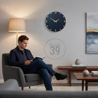 A navy blue wall clock with gold-colored hands and Roman numerals. The clock is round and mounted on a light gray wall. The image also shows a person reading a book in a gray armchair.