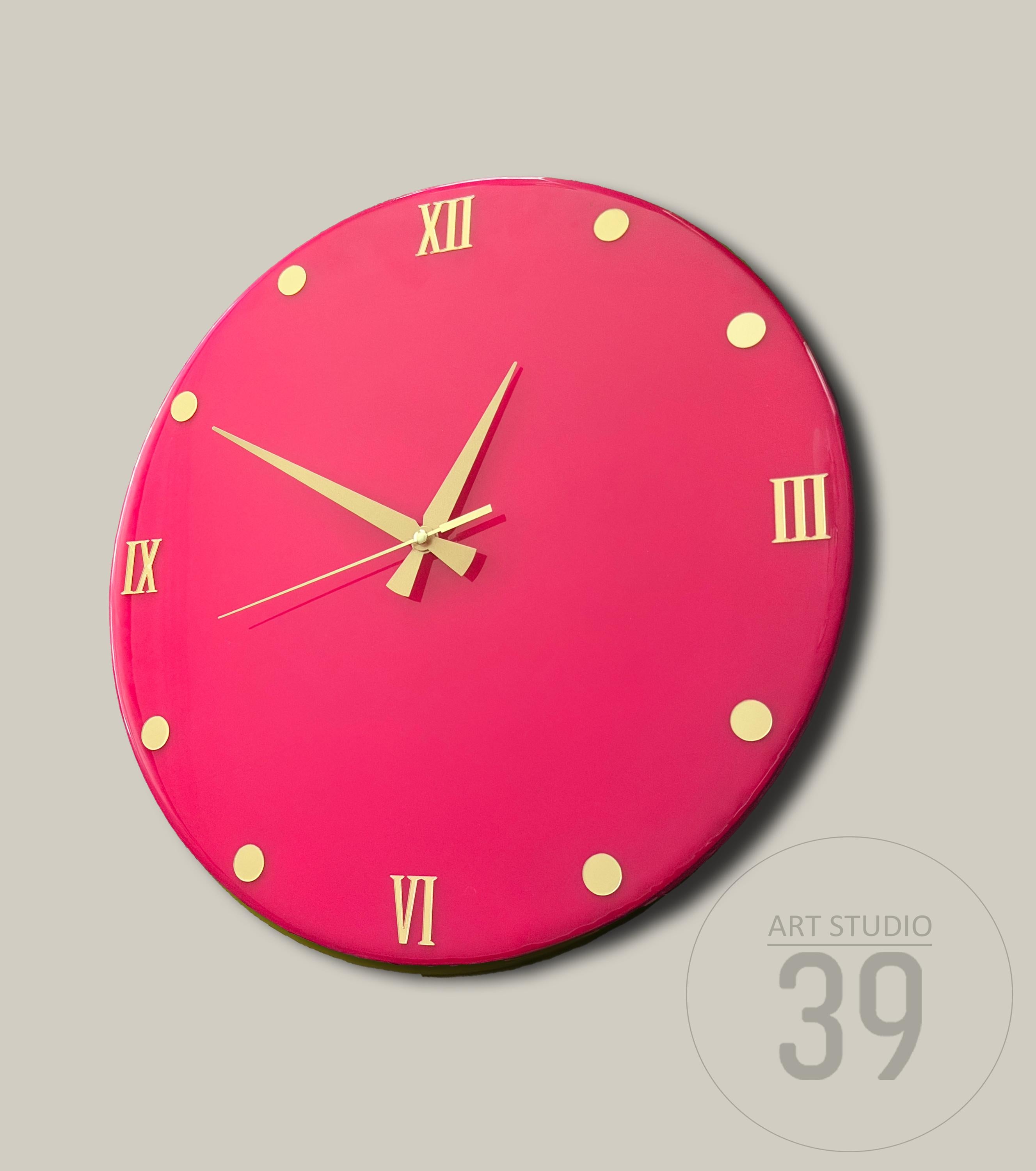 A round, hot pink wall clock with gold-colored hands and Roman numeral hour markers. The clock face has gold-colored dots marking the hours. The clock is from Art Studio 39.