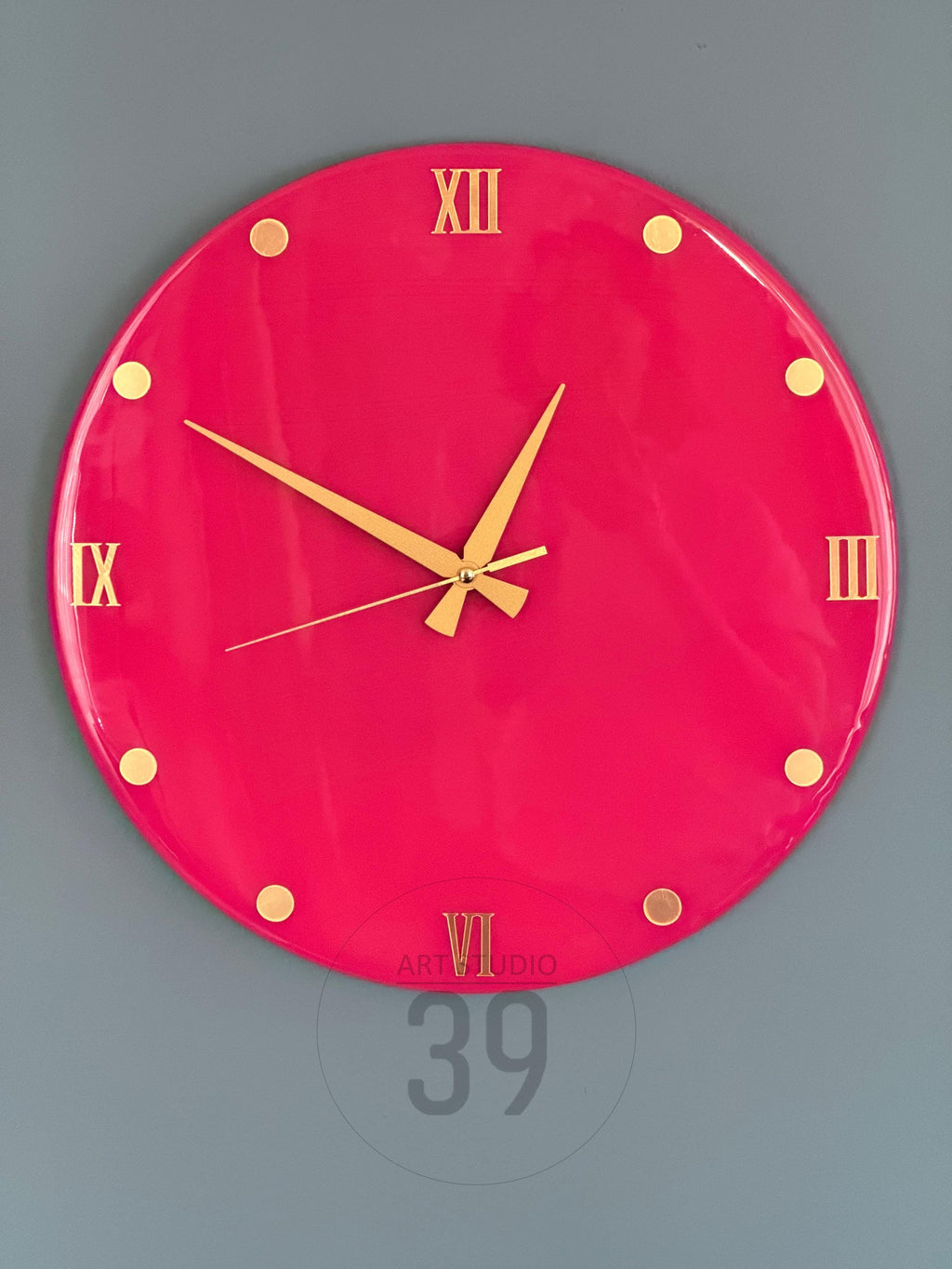A round, hot pink wall clock with gold-colored hands and Roman numeral hour markers. The clock has gold-colored dots marking the hours and the text "ART STUDIO 39" is visible at the bottom.