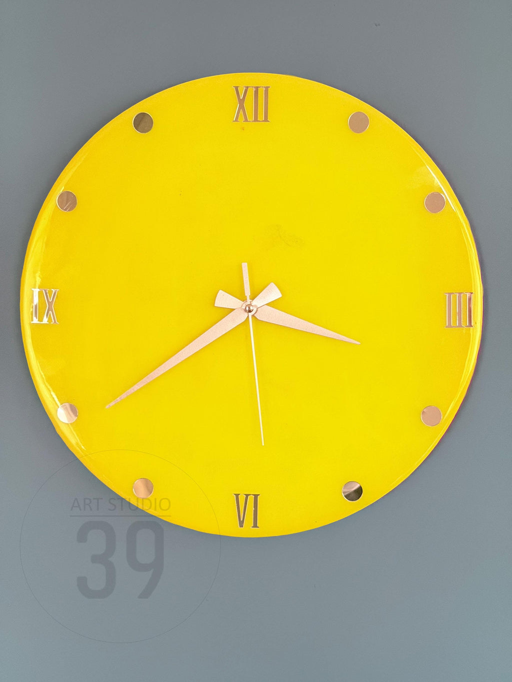 A round, bright yellow wall clock with gold-colored Roman numerals and hands. The clock face has gold-colored dots marking the hours. The clock reads 6:10. The text "ART STUDIO 39" is visible on the bottom left.