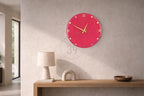 A round, hot pink wall clock with gold-colored hands and Roman numerals. The clock is mounted on a light gray wall. The image also shows a table with a lamp, a decorative arch, and a wooden bowl.