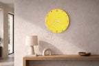 A bright yellow wall clock with Roman numerals and hands. The clock is mounted on a light gray wall. A wooden shelf below holds a lamp, a decorative arch, and a wooden bowl.