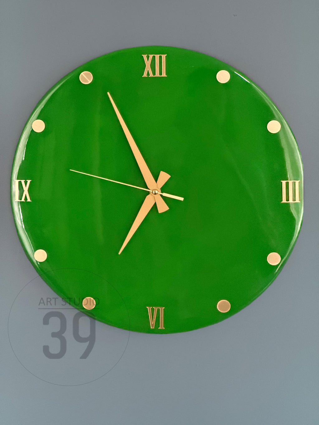 A round, bright green wall clock with gold-colored Roman numerals and hands. The clock face has gold-colored dots marking the hours. The clock is mounted on a gray wall. The clock's diameter is approximately 12 inches.