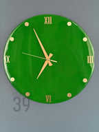 A round, bright green wall clock with gold-colored Roman numerals and hands. The clock face has gold-colored dots marking the hours. The clock is mounted on a gray wall. The clock's diameter is approximately 12 inches.