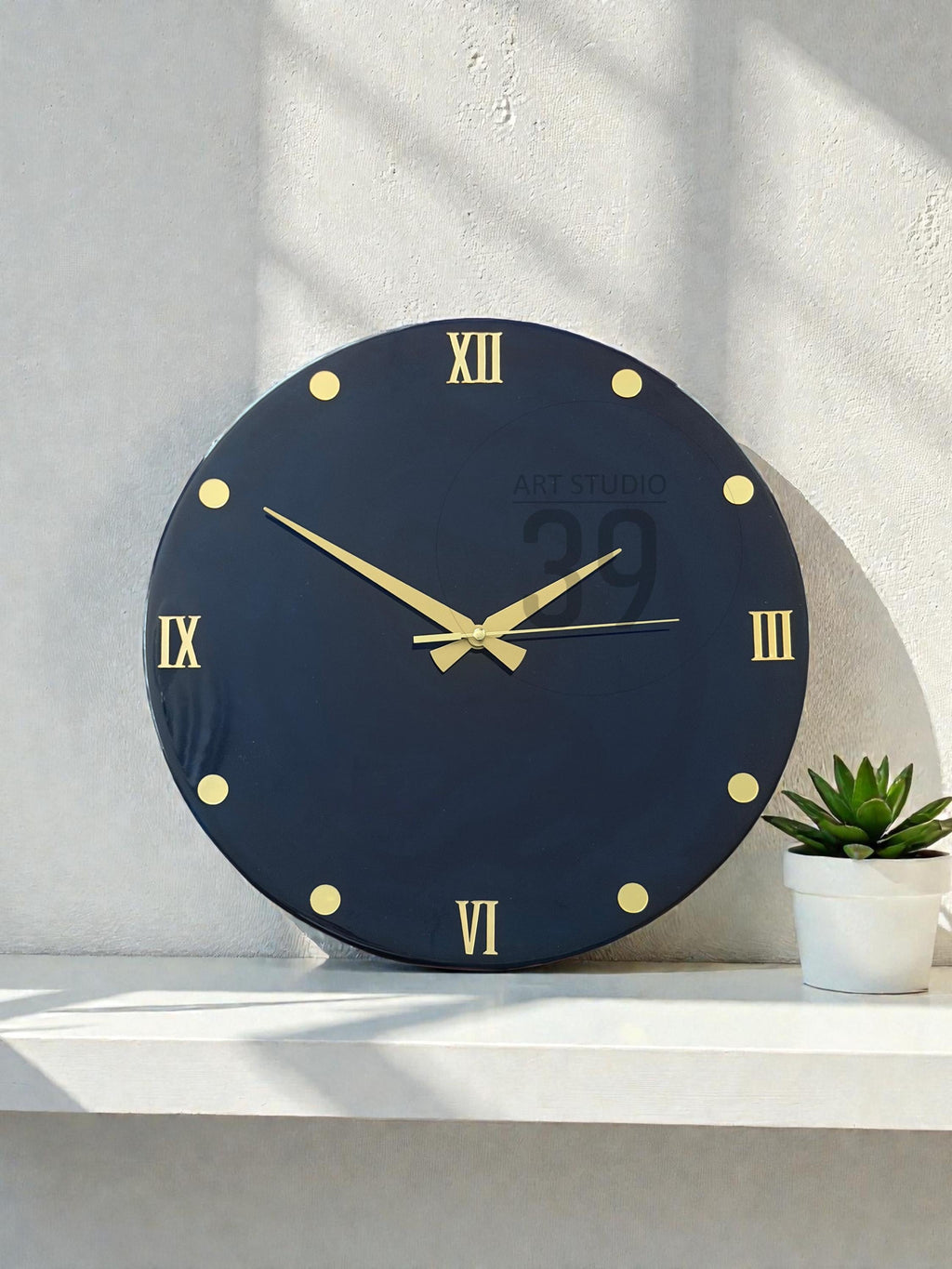 A round, navy blue wall clock with gold-colored Roman numerals and hands. The clock face has gold dots marking the hours. The text "ART STUDIO 29" is printed on the clock face. A small potted succulent sits to the right of the clock.