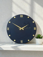 A round, navy blue wall clock with gold-colored Roman numerals and hands. The clock face has gold dots marking the hours. The text "ART STUDIO 29" is printed on the clock face. A small potted succulent sits to the right of the clock.