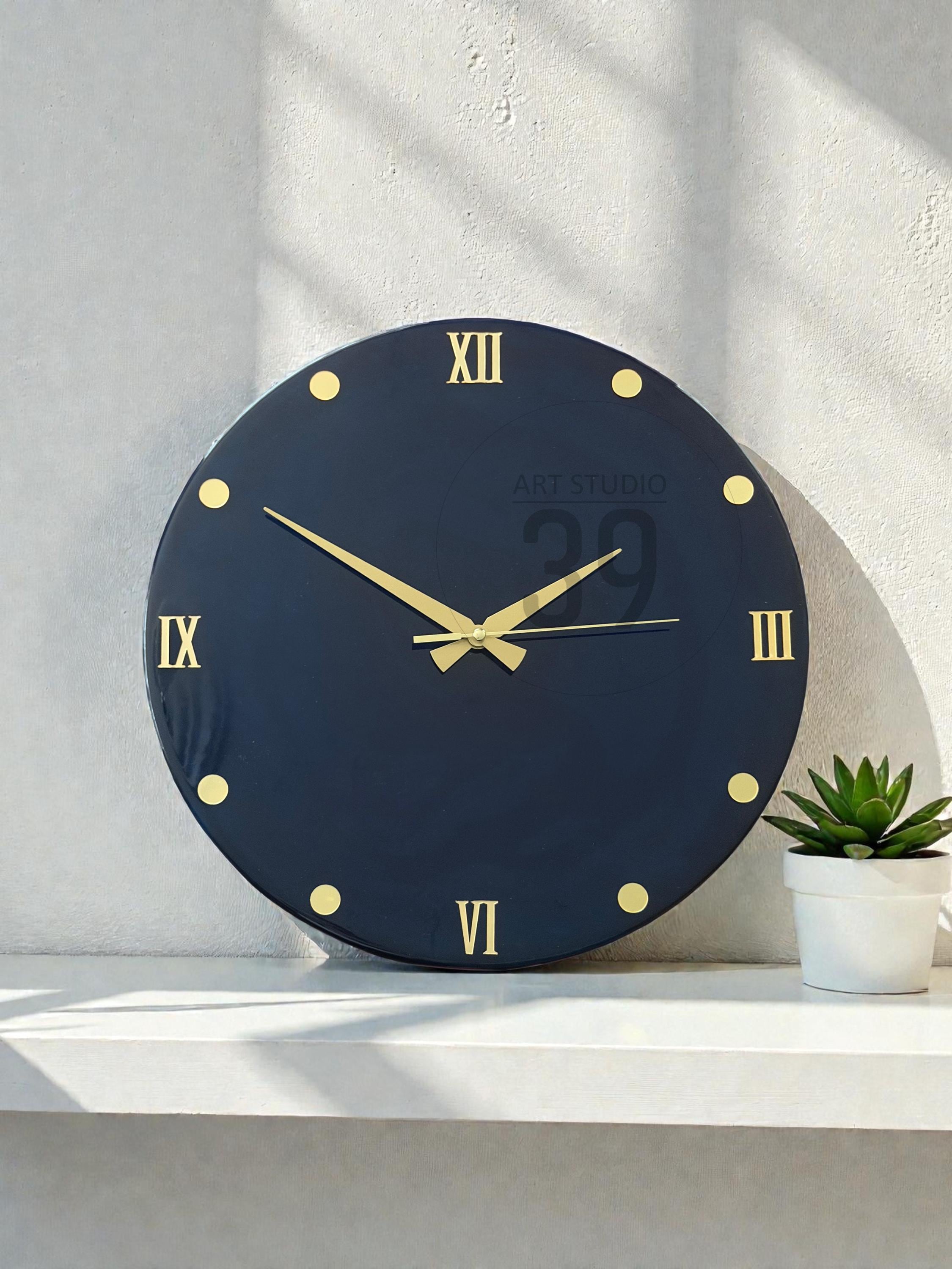 A round, navy blue wall clock with gold-colored Roman numerals and hands. The clock face has gold dots marking the hours. The text "ART STUDIO 29" is printed on the clock face. A small potted succulent sits to the right of the clock.