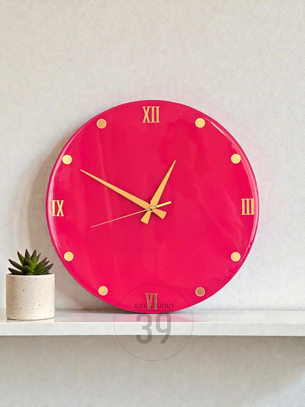 A round, hot pink wall clock with gold-colored Roman numerals and hands. The clock has gold dot markers and the text "ART STUDIO 39" at the bottom. A small potted succulent sits on a white shelf below the clock.