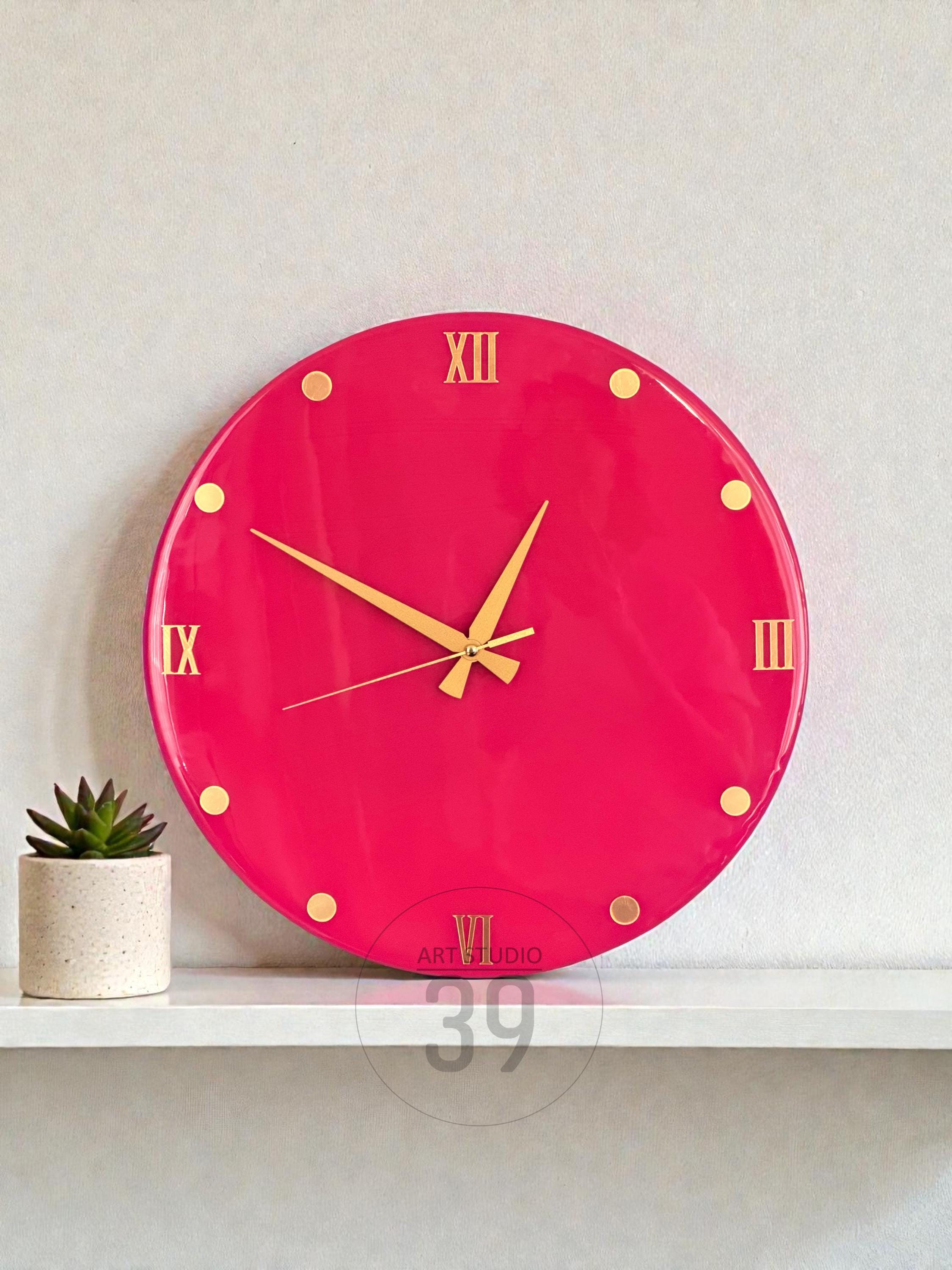 A round, hot pink wall clock with gold-colored Roman numerals and hands. The clock has gold dot markers and the text "ART STUDIO 39" at the bottom. A small potted succulent sits on a white shelf below the clock.