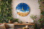 A round wall clock with a beach scene design. The top half features a blue ocean with white waves, while the bottom half depicts a sandy beach. Gold Roman numerals mark the hours. The clock hands are gold.