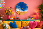 A round wall clock with a blue ocean design, gold hands, and Roman numerals. The clock is mounted on a pink wall above a teal cabinet. The room is decorated with flowers, a lamp, and a yellow sofa.