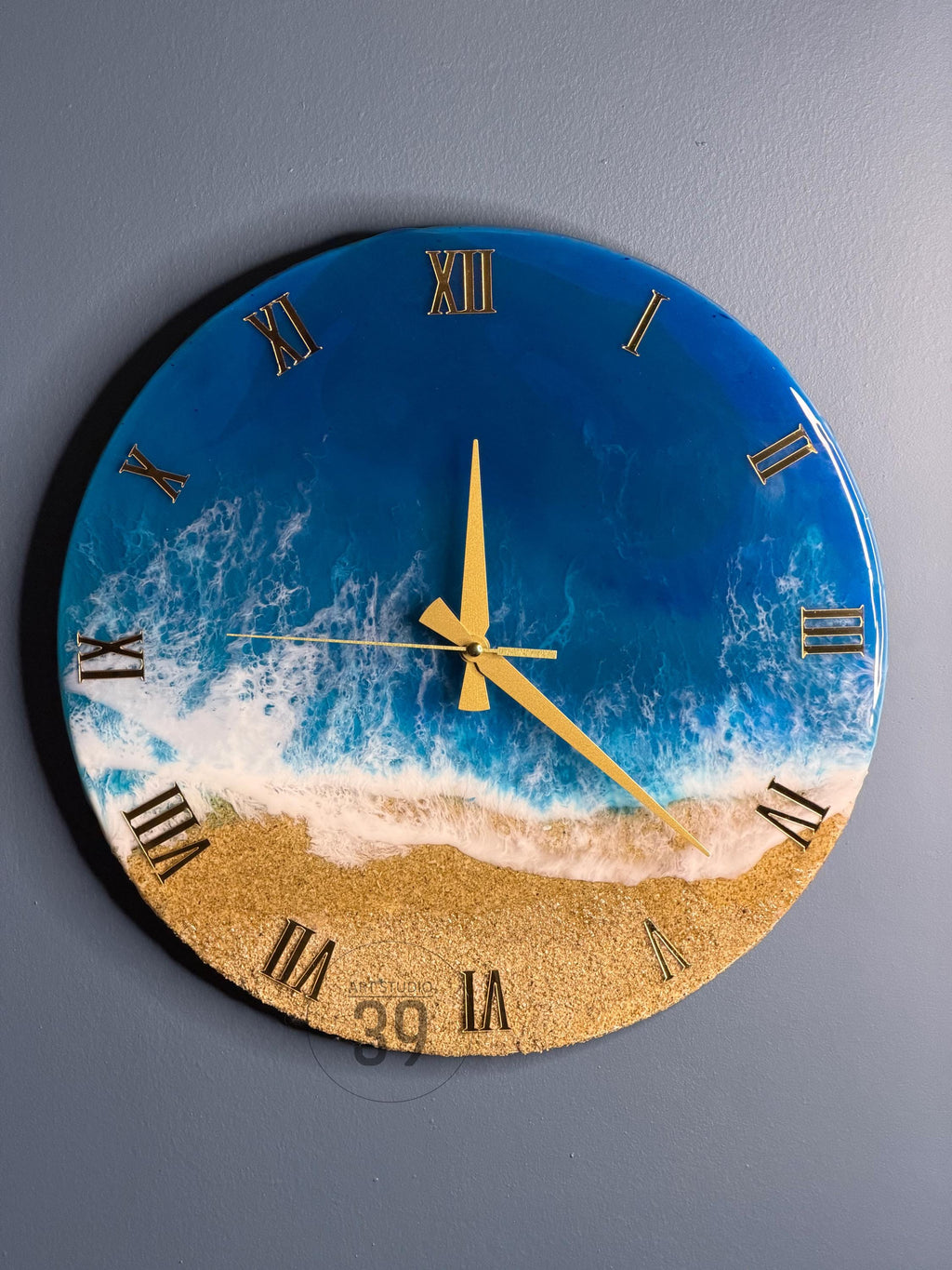 Round wall clock with a beach scene design. The clock face features a blue ocean, white foamy waves, and a sandy beach. Gold Roman numerals mark the hours, and gold hands indicate the time. The clock is a decorative item.