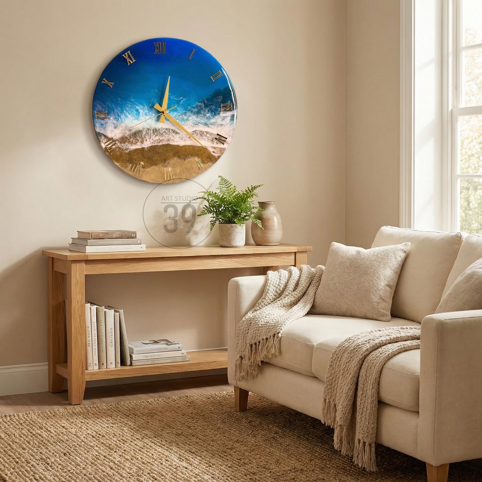 A round wall clock with a beach scene design. The clock face features a blue and white ocean design with gold Roman numerals and hands. The clock is hanging on a beige wall above a wooden console table.