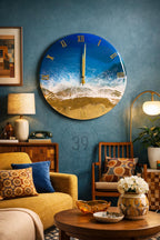 A large, round wall clock with a beach-themed design. The clock face features a blue ocean and sandy beach, with gold Roman numerals and hands. The clock is hanging on a blue wall in a living room setting.