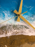 A round wall clock with a beach scene design. The clock face features a blue ocean, white foamy waves, and a sandy beach. Gold-colored clock hands point to the time. The number 39 is visible on the clock face.
