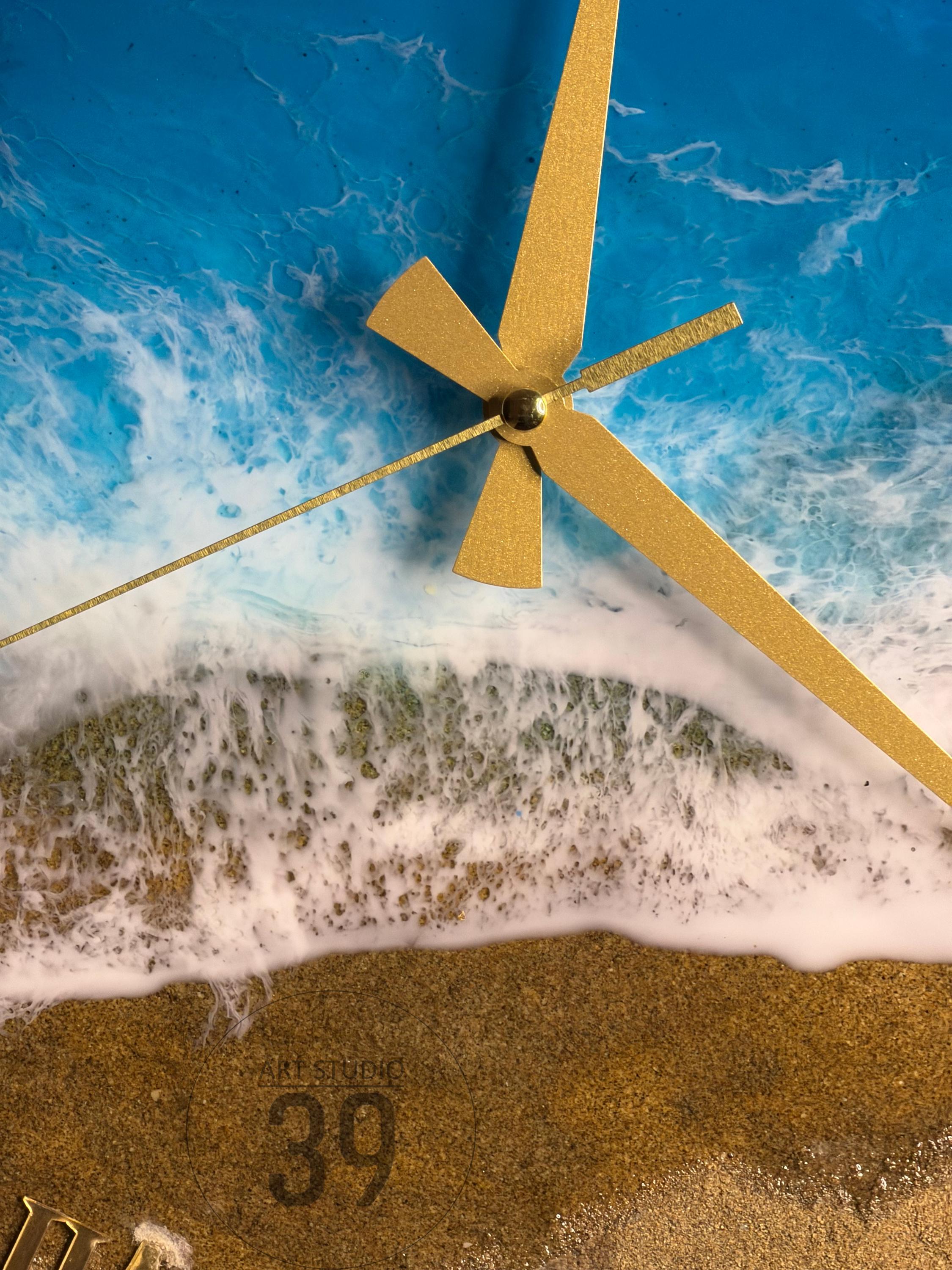 A round wall clock with a beach scene design. The clock face features a blue ocean, white foamy waves, and a sandy beach. Gold-colored clock hands point to the time. The number 39 is visible on the clock face.