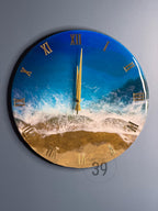 Round wall clock with a beach scene design. The clock face features a blue ocean, white waves, and golden sand. Gold Roman numerals mark the hours. The clock hands are gold.