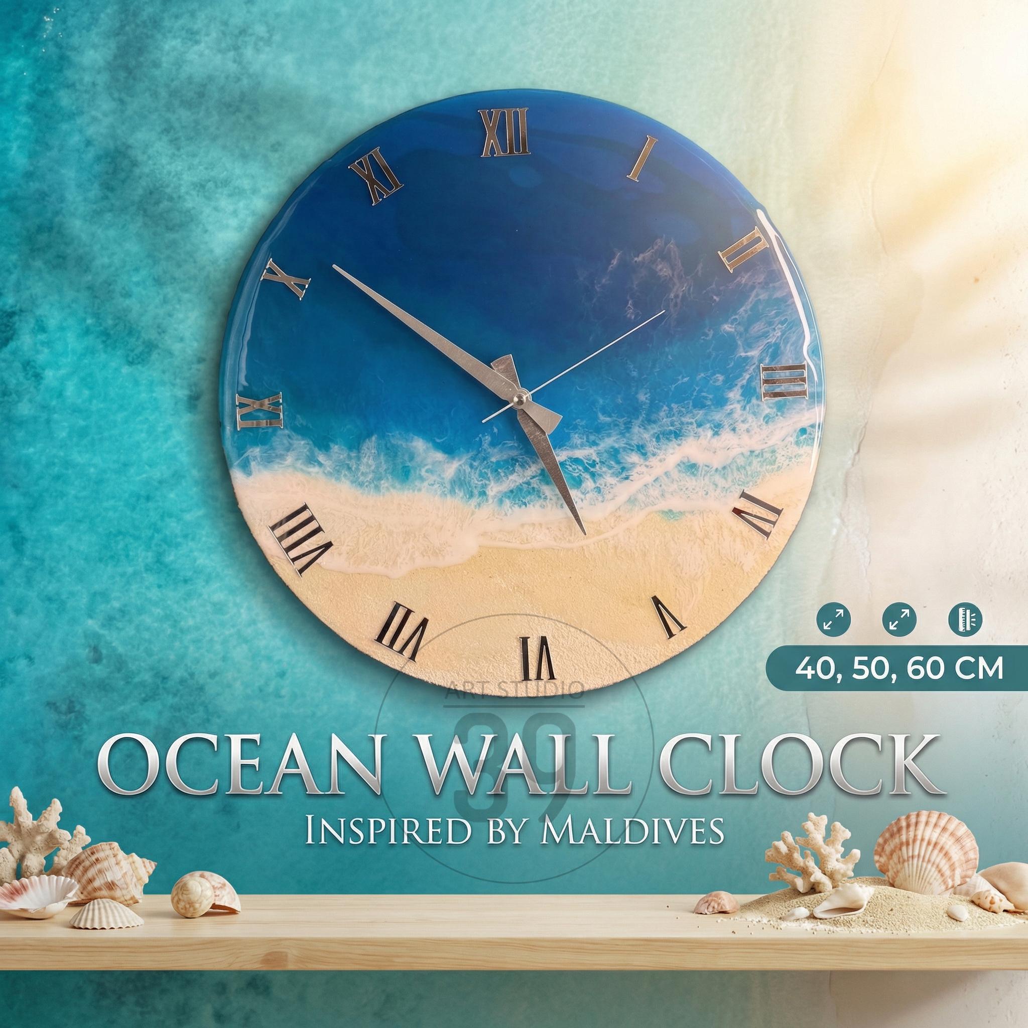 Round wall clock with a beach and ocean design. The clock face features a blue ocean, white waves, and a sandy beach. Roman numerals mark the hours. The clock is available in 40, 50, and 60 cm sizes. The text "OCEAN WALL CLOCK INSPIRED BY MALDIVES" is displayed.