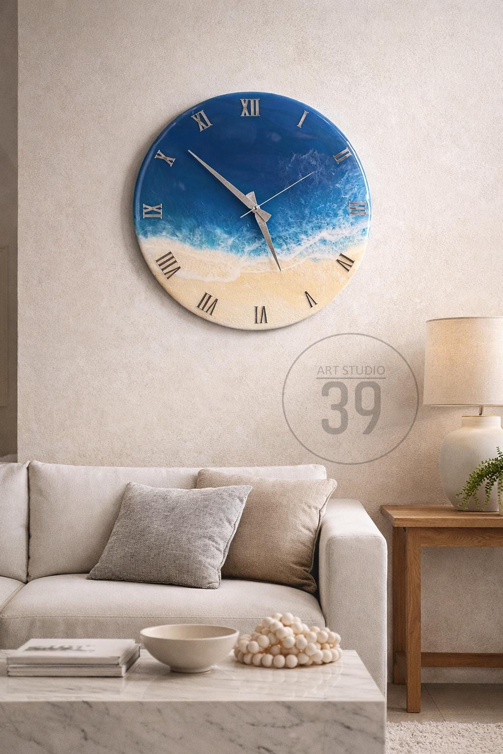A round wall clock with a blue and white ocean-themed design. The clock face features Roman numerals and silver hands. The clock is mounted on a light-colored wall above a sofa and coffee table.