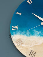 A round wall clock with a beach-themed design. The clock face features a gradient of blue and white, resembling the ocean and waves, with a sandy beach at the bottom. Silver Roman numerals mark the hours. The clock hands are silver.