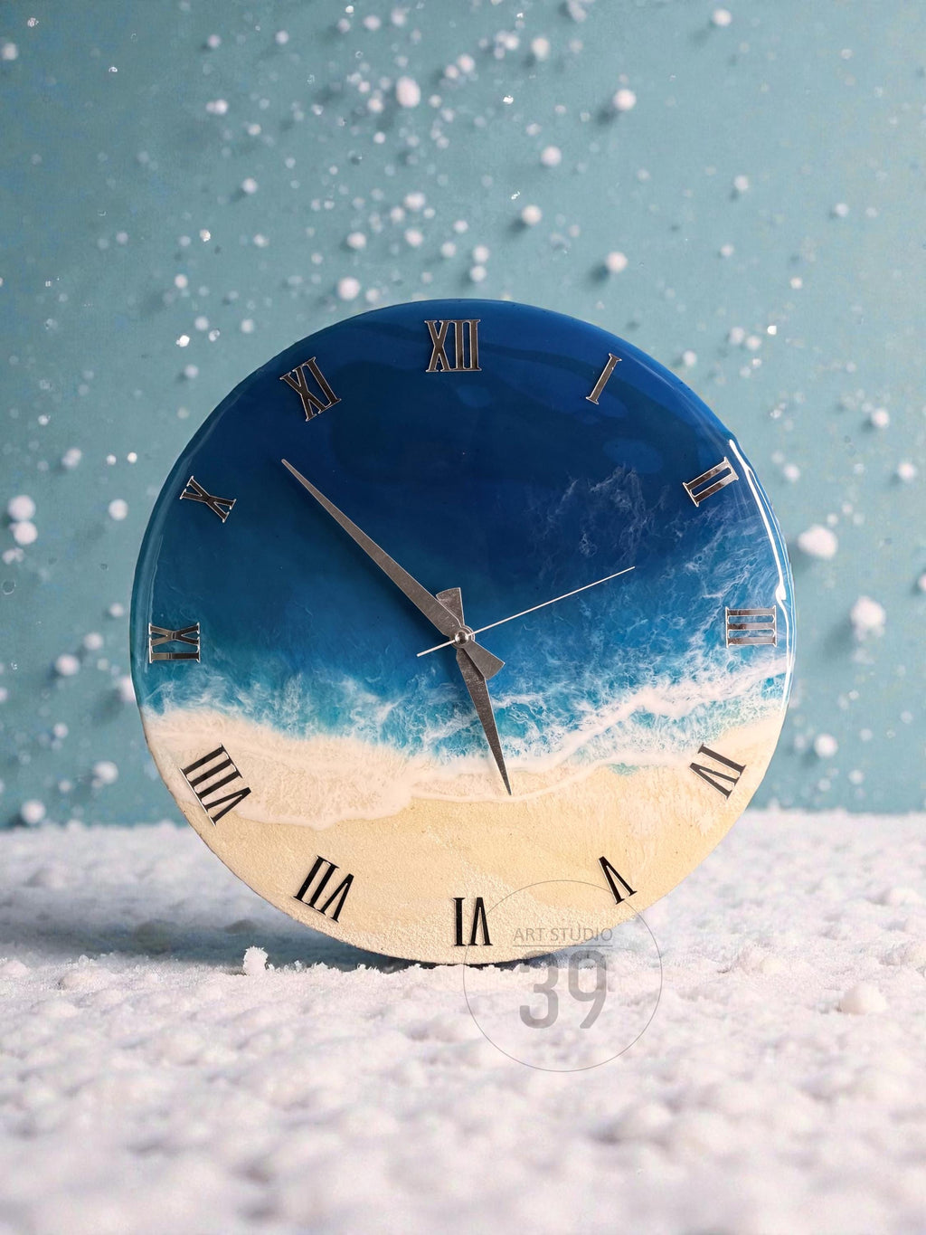 Round wall clock with a blue and white ocean-themed design. The clock face features Roman numerals and silver hands. The bottom half of the clock face is a sandy color, and the top half is blue.