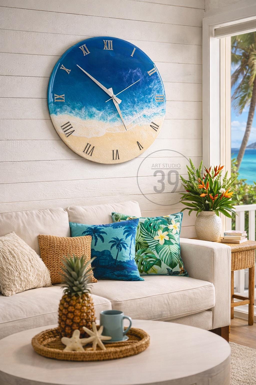 A large, round wall clock with a beach-themed design. The clock face features a blue ocean and sandy beach, with silver hands and Roman numerals. The clock is mounted on a white wall above a white sofa with decorative pillows.