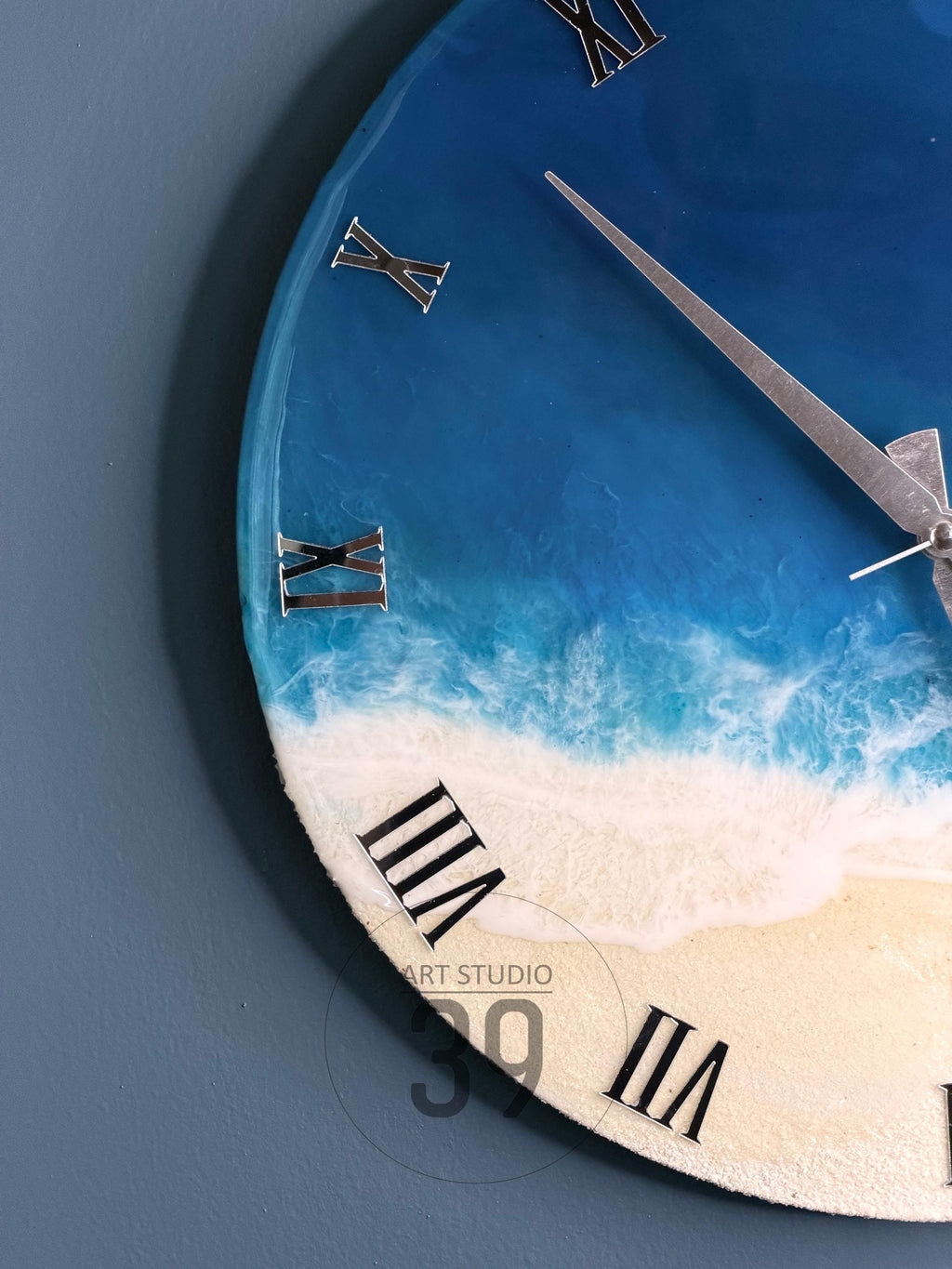 Round wall clock with a beach-themed design. The clock face features a blue and white ocean scene with silver Roman numerals and clock hands. The clock has the text "ART STUDIO 39".