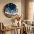 A round wall clock with a coastal design. The clock face features a dark blue ocean scene with white waves and a sandy beach. Silver Roman numerals mark the hours. The clock is mounted on a beige wall.
