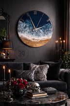 A large, round wall clock with a beach-themed design. The clock face features a dark blue sky, white waves, and a golden sand beach. Silver hands and Roman numerals complete the design. The clock is mounted on a dark gray wall.