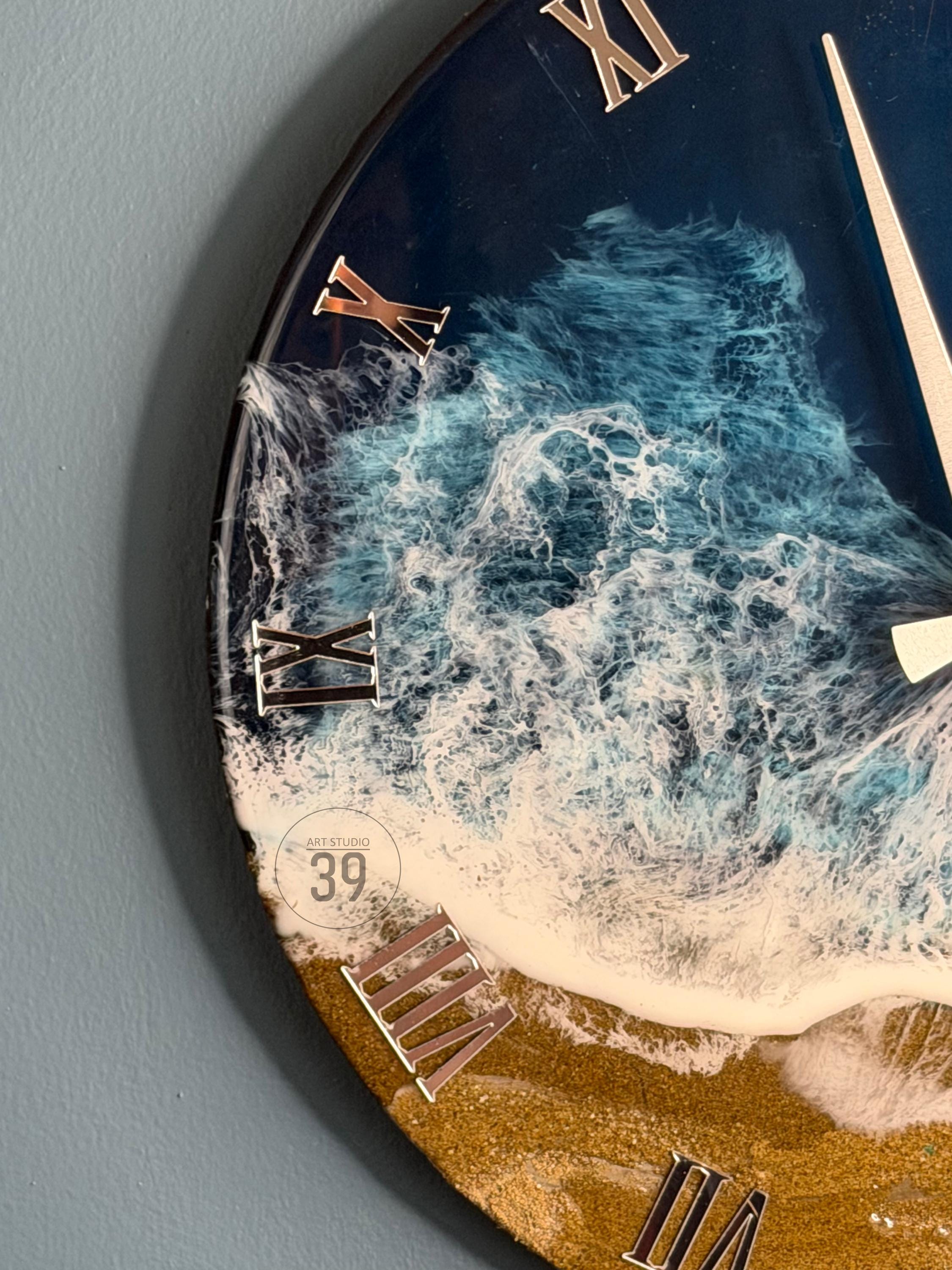 Round wall clock with a resin ocean wave design. The clock face features a dark blue background with white foamy waves, a sandy beach, and metallic Roman numerals. The clock hands are silver, and the clock has the text "ART STUDIO 39".