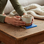 A speckled ceramic mug with a brown base, filled with a hot beverage, rests on a blue coaster. Steam rises from the mug, suggesting warmth. The mug is held over a wooden table.