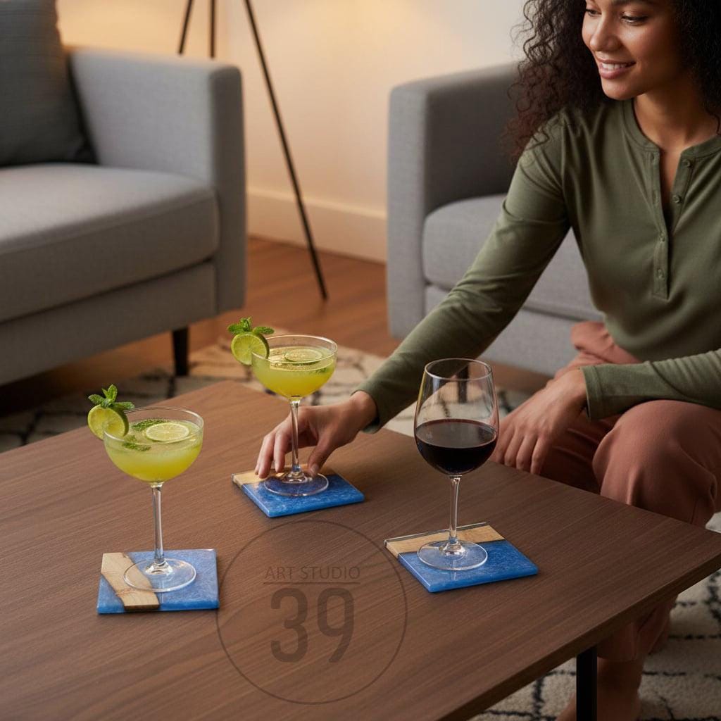 A woman places a cocktail glass on a blue and gold square coaster. Two other cocktails and a glass of red wine sit on similar coasters on a brown table. The coasters have a blue marbled design. The text "ART STUDIO 39" is in the image.
