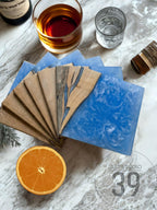 A collection of wooden coasters with blue resin accents, fanned out on a marble surface. The coasters are paired with a glass of amber liquid, a glass of water, and a sliced orange. The image also includes the text "ART STUDIO 39".