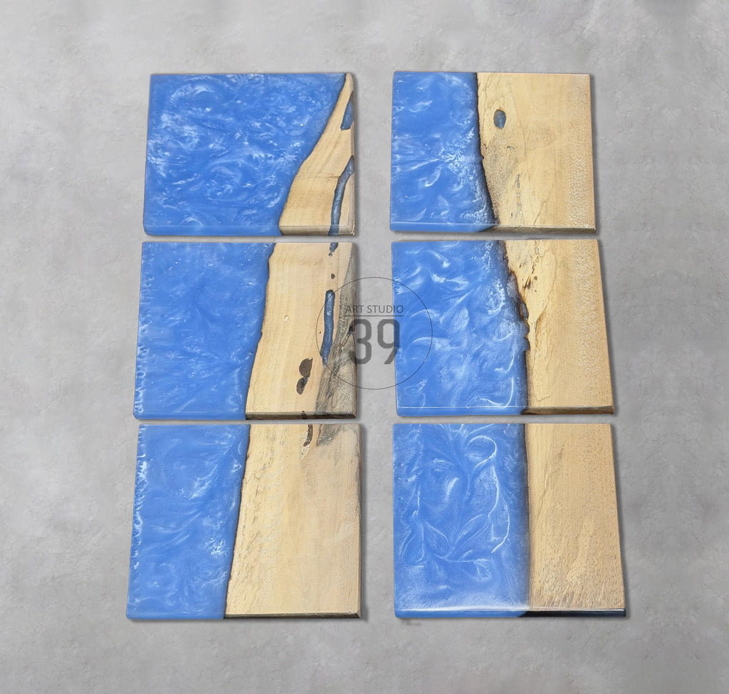 Six square coasters with a unique design. Each coaster features a combination of light blue resin and natural wood. The resin has a textured appearance, and the wood has a light brown color. The coasters are arranged on a gray surface.