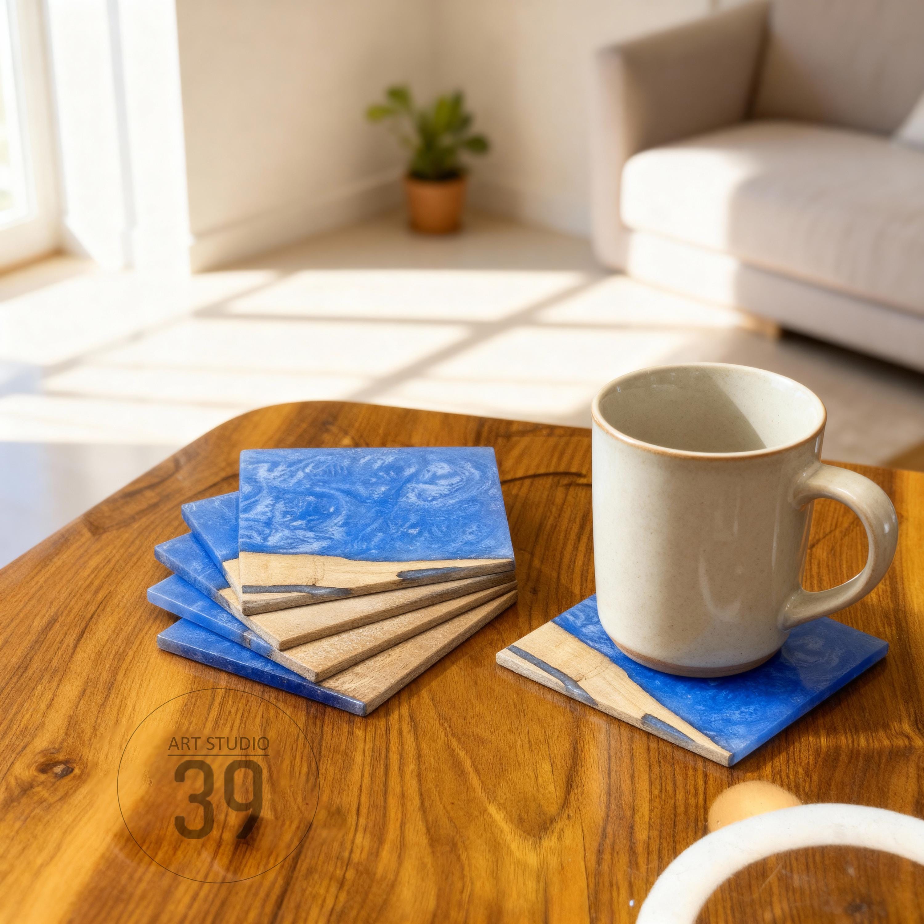 A set of square coasters with a blue resin top and a wooden base. A beige mug sits on one coaster. The coasters are on a wooden table. The text "ART STUDIO 39" is visible.