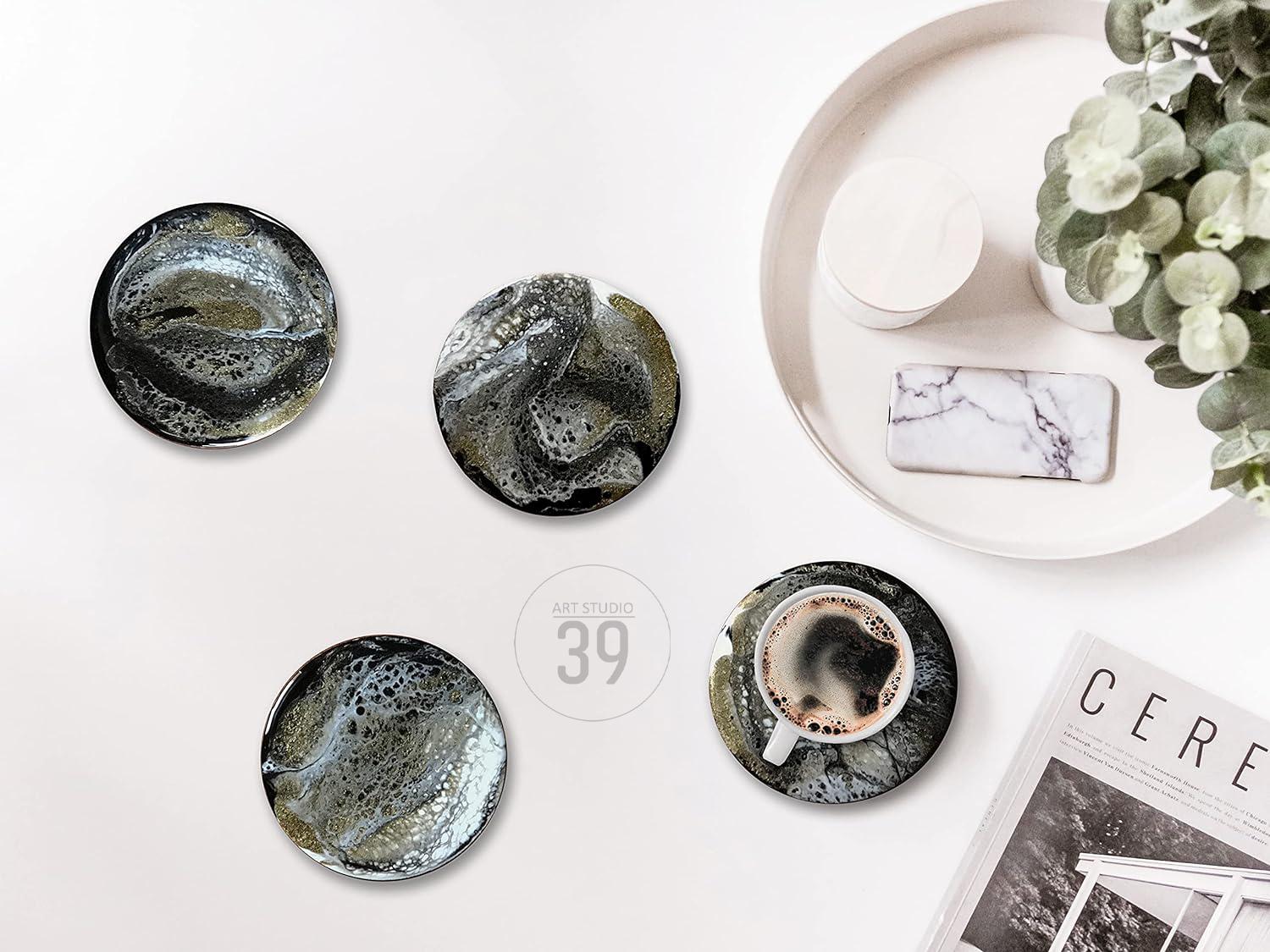 Four round coasters with a black, gold, and white abstract design. One coaster holds a white coffee cup filled with coffee. A white tray with a marble phone stand and a small white container is in the background.