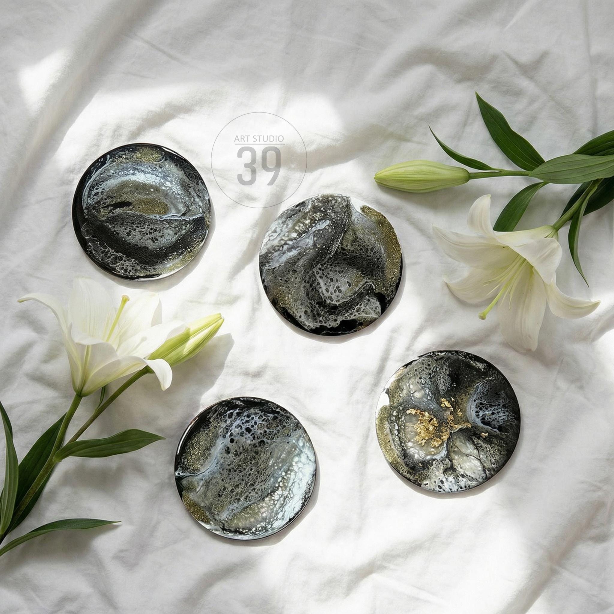 Five round coasters with abstract designs in black, gold, and white, arranged on a white surface. The coasters have a glossy finish and are surrounded by white lilies. The text "ART STUDIO 39" is visible.