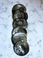 A set of six round coasters with a marbled design in black, gold, and white. The coasters have a glossy finish and are arranged in a vertical line. The background is a white marble surface. The text "STUDIO 39" is visible.