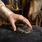 A gold-toned ring with a floral design and a smaller ring with small stones adorn a hand. The hand is touching a round, black and gold decorative object. The object has a unique, marbled appearance. The background is blurred.