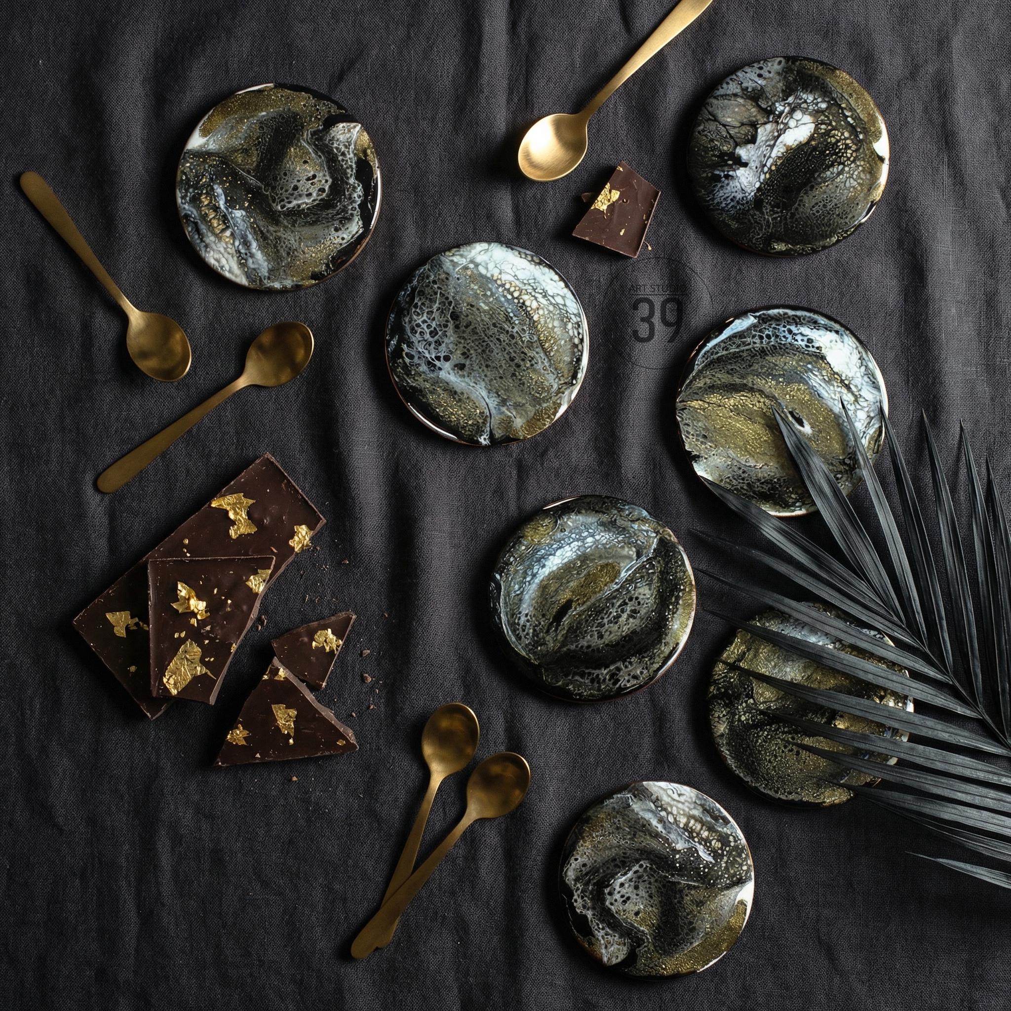 An overhead shot features several round coasters with a black, white, and gold marbled design. Gold spoons and pieces of chocolate with gold leaf accents are scattered around the coasters. The number 39 is visible.