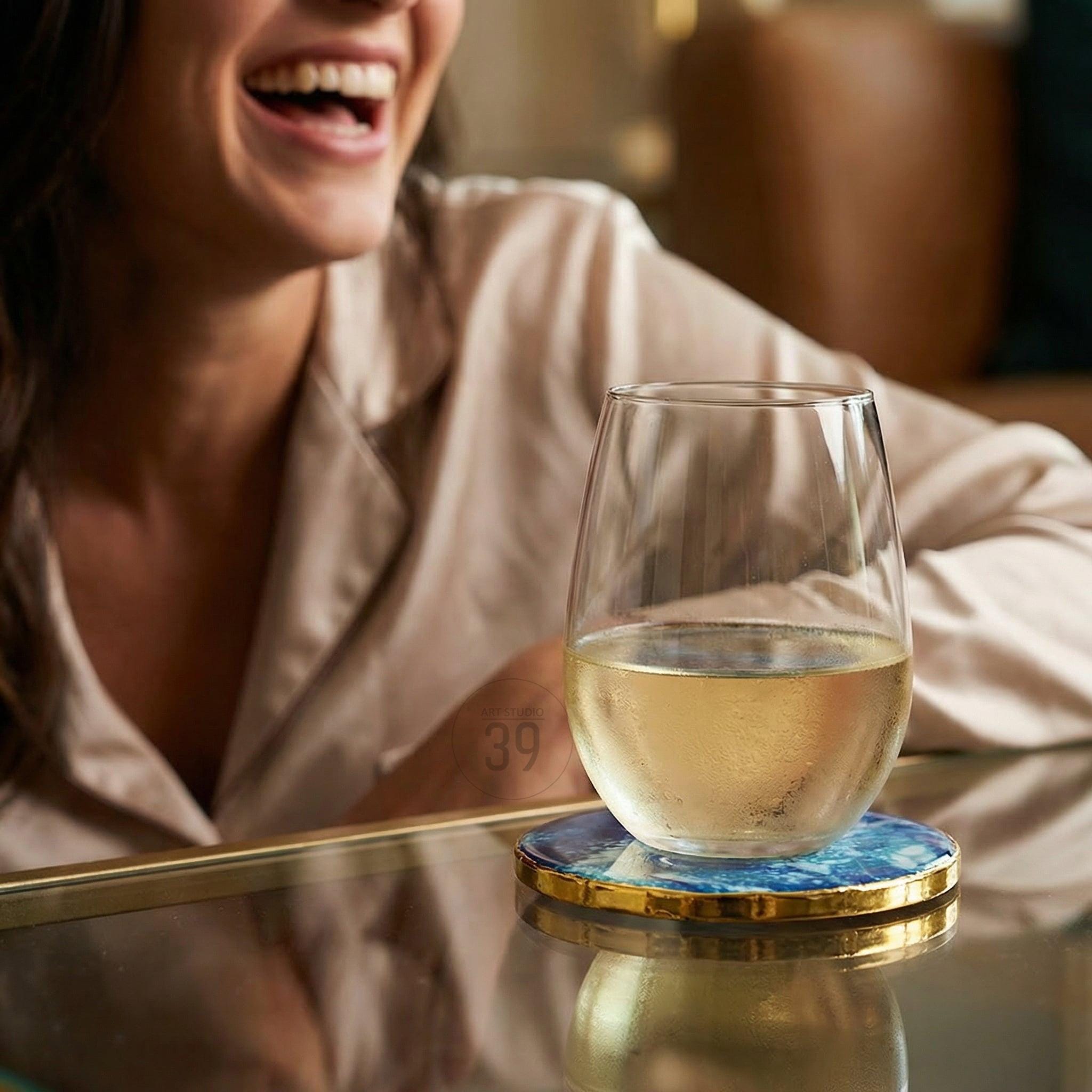 A stemless wine glass filled with white wine sits on a blue and gold coaster. The coaster has a marbled blue design with a gold-colored edge. The glass is on a reflective surface.