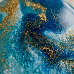 Abstract artwork with a fluid design in shades of blue and white, accented with gold glitter. The piece has a glossy finish, creating a dynamic and textured appearance. The artwork evokes a sense of the ocean.