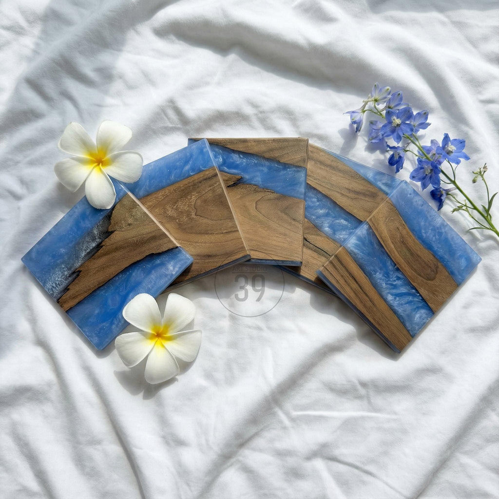 Set of four square coasters with a unique design. Each coaster features a combination of wood and blue resin. The wood has a natural grain, and the resin has a marbled effect. White flowers and blue flowers are arranged around the coasters.