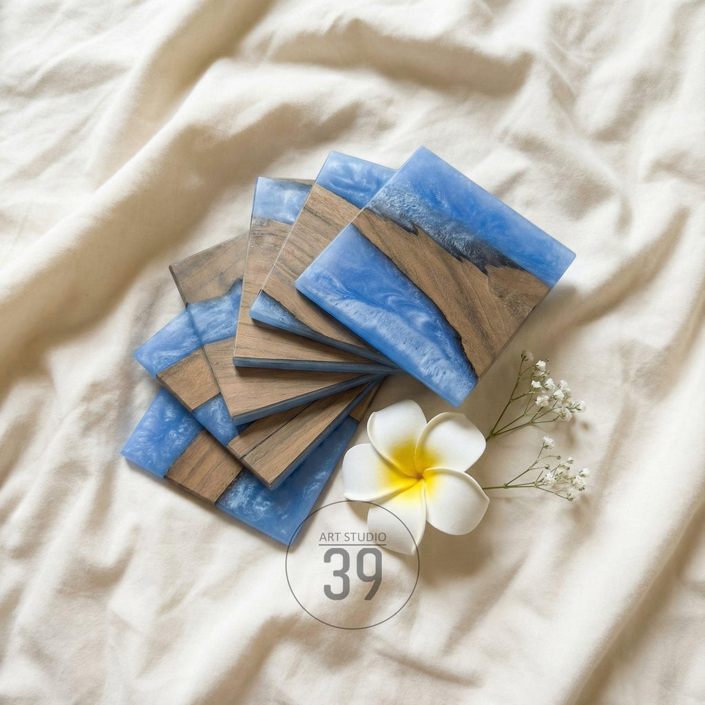 A stack of square coasters with a blue resin design and wood grain accents. The coasters are arranged on a white surface, with a white flower and the text "ART STUDIO 39" in the corner.