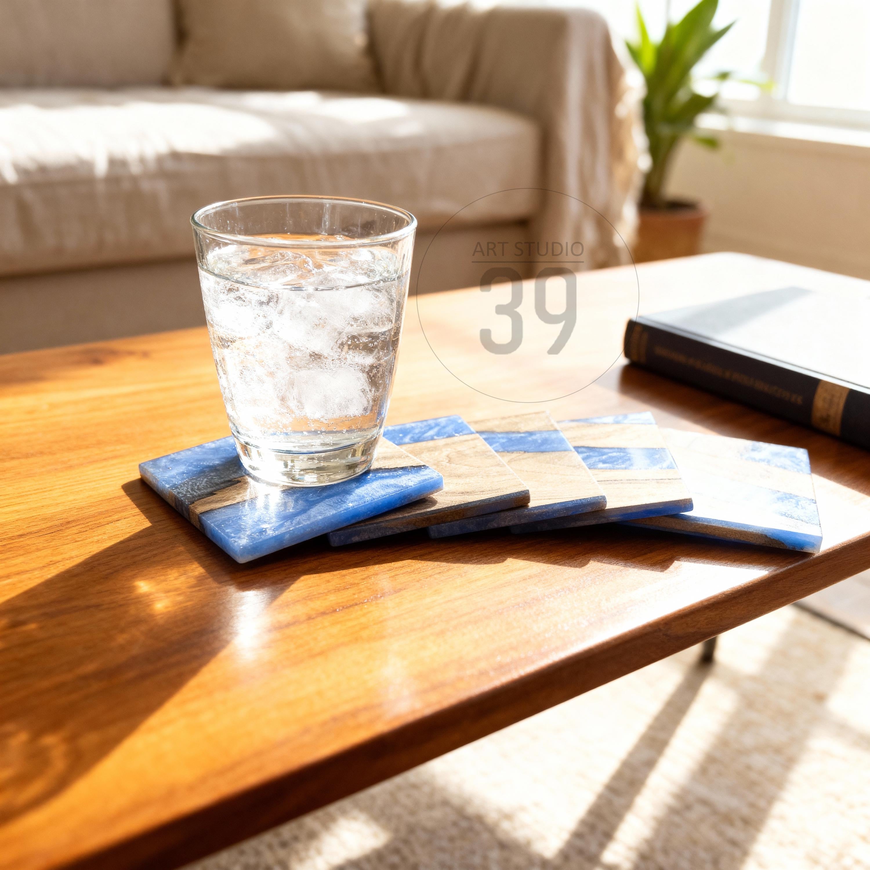 A clear glass of water with ice sits on a set of square coasters. The coasters are made of wood and blue resin. The coasters are on a wooden table. The image has the text "ART STUDIO 39" in the background.