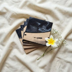 A stack of square wooden coasters with a black resin top. The coasters have a natural wood grain finish on the bottom half. A white flower with a yellow center and small white flowers are placed near the coasters. The text "ART STUDIO 20" is visible.