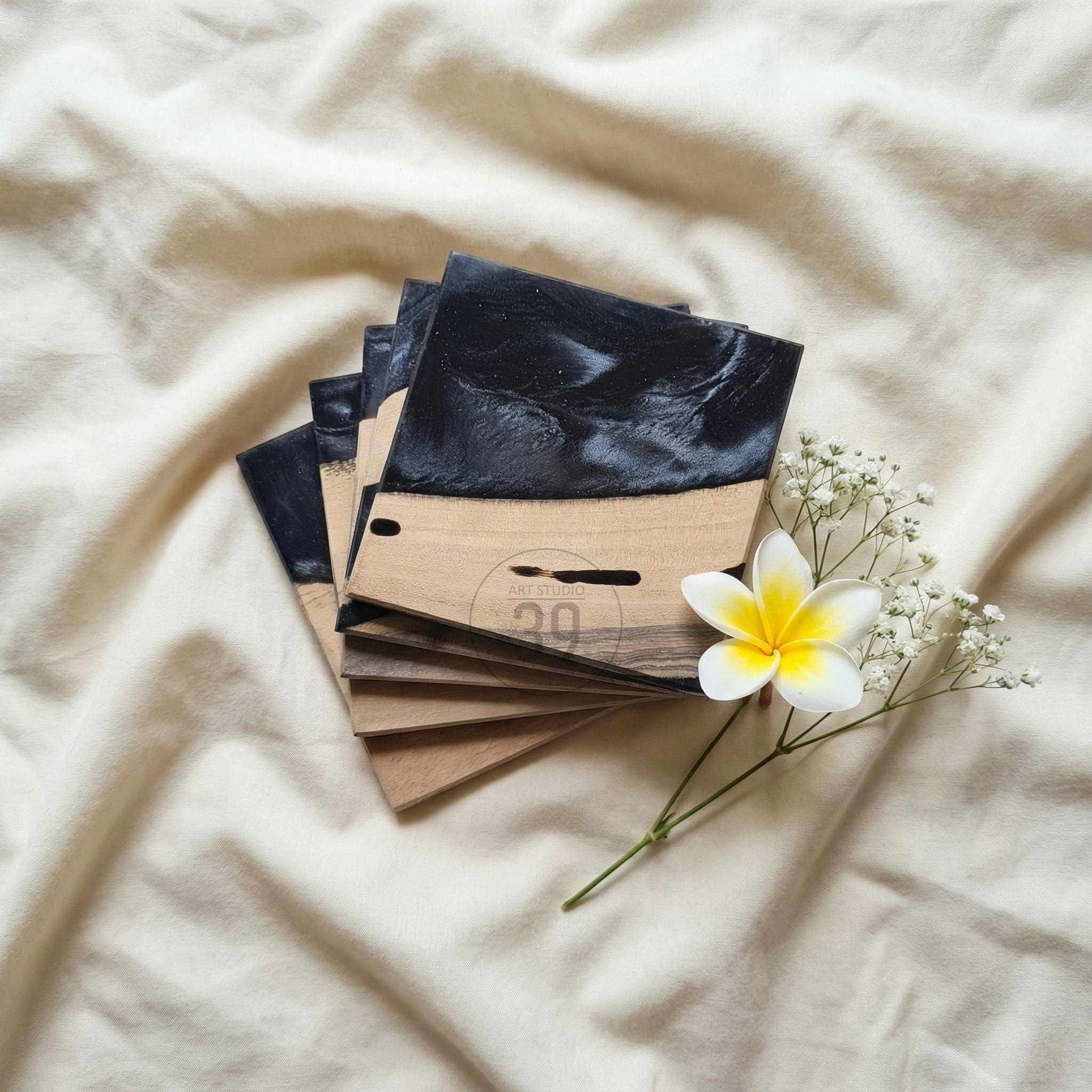 A stack of square wooden coasters with a black resin top. The coasters have a natural wood grain finish on the bottom half. A white flower with a yellow center and small white flowers are placed near the coasters. The text "ART STUDIO 20" is visible.