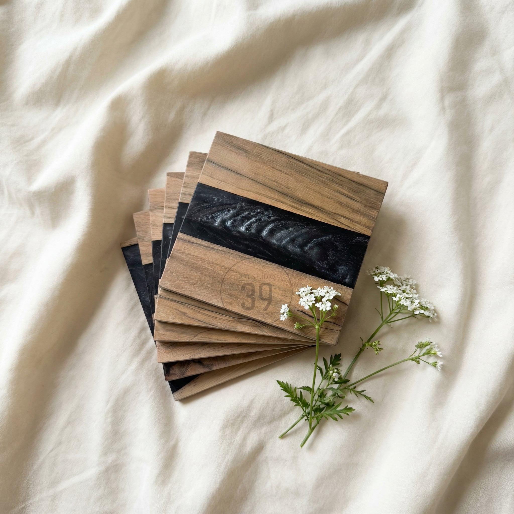 A stack of square wooden coasters with a dark, black resin inlay. The coasters have a natural wood grain pattern and the text "39" is visible. A sprig of white flowers rests beside the coasters.